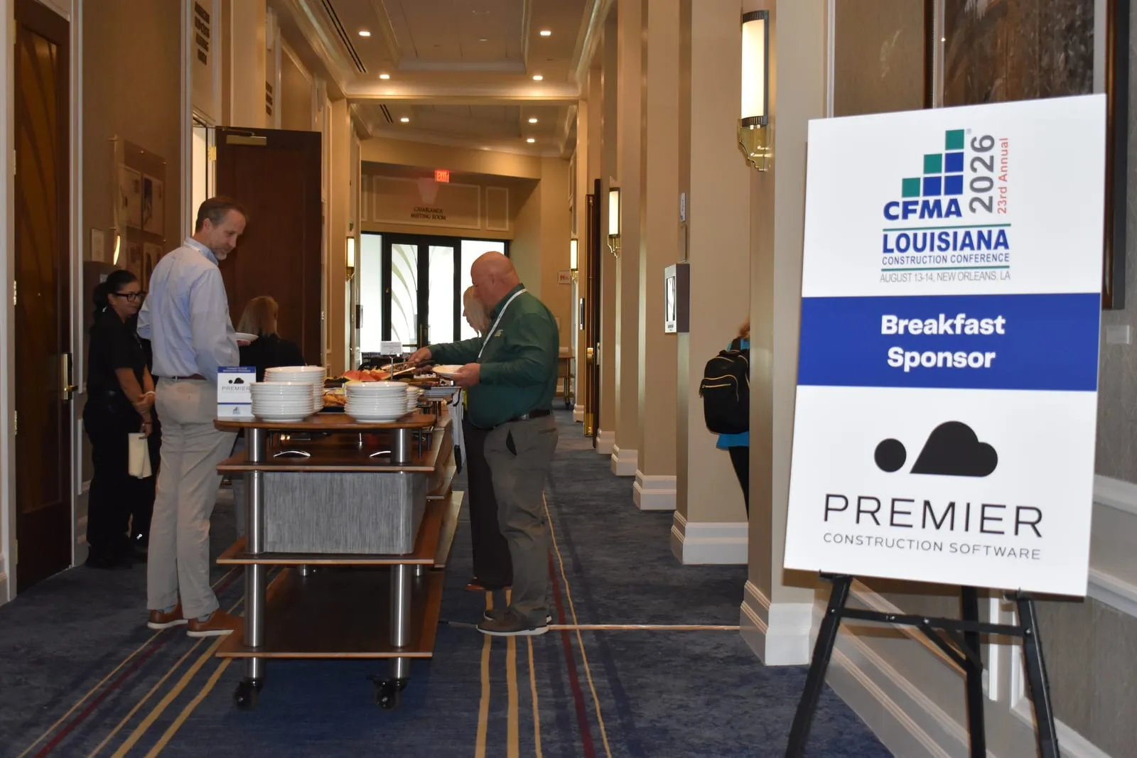 A breakfast sponsor sign for the 23rd Annual CFMA Louisiana Construction Conference, August 13-14 in New Orleans, standing beside a buffet where attendees are serving themselves and talking.